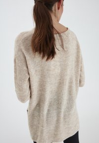 Pull oversized beige avec un tricot texturé, un ourlet côtelé et des épaules tombantes. Présente une apparence douce et légèrement transparente, avec des fentes sur les côtés.