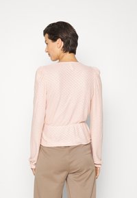 Top de manga longa rosa claro com padrão texturizado, mangas bufantes franzidas e cintura peplum. Combinado com calças bege, mostrado de costas.