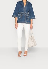 Jeansowa kurtka z szerokimi rękawami i paskiem, noszona na białej bluzce i dopasowanych białych spodniach, w połączeniu z jasną torebką i szpilkami.