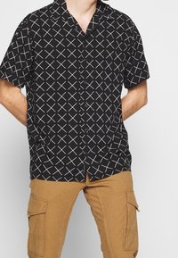 Kurzarm schwarzes Shirt mit einem weißen X-Muster, klassischem Kragen und Knöpfen an der Vorderseite. Kombiniert mit hellbraunen Cargo-Hosen.