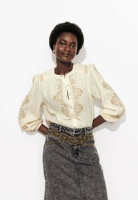 Blouse crème avec des manches bouffantes et des broderies florales dorées, associée à une jupe en denim taille haute avec une ceinture brodée.