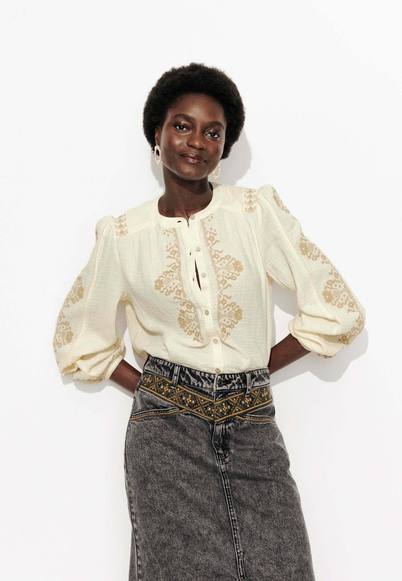 Blouse crème avec des manches bouffantes et des broderies florales dorées, associée à une jupe en denim taille haute avec une ceinture brodée.