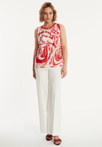 Mouwloze top met een rood en roze abstract patroon, gerimpelde halslijn en decoratieve accenten. Gepaard met witte wijde pijpenbroek en rode sandalen.