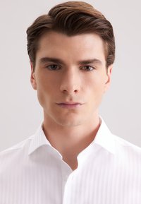 Wit gestreepte shirt met een geknoopte kraag, met een gladde textuur. Close-up van het gezicht van een mannelijke model, neutrale uitdrukking, bruin haar.