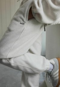 Heledad hallhalli pehmete tekstuuriga dressipüksid, millel on "self." logo, elastsed varrukad ja lukuga taskudetail. Kandmine hallide vestidega.