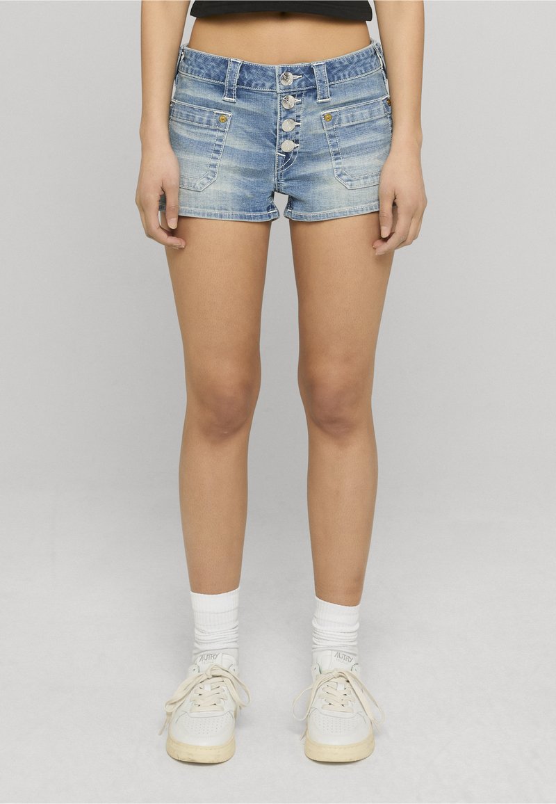 Shorts en jean bleu clair avec un ourlet effiloché, une fermeture devant à cinq boutons et deux poches latérales. Assortis de baskets blanches et de chaussettes.