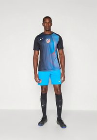 Mees seisab, kandes musta ja sinist Atlético Madridi jalgpalliriietust koos mustade sokkide ja ketsidega lihtsa taustaga.