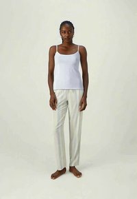 Moteris stovinti basomis kojomis, vilkinti baltą be rankovių marškinėlį ir šviesius dryžuotus kelnes prieš paprastą šviesų fono.