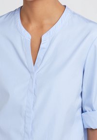 Jasnoniebieska koszula z kołnierzykiem, o dekolcie z wcięciem, krótkich, podwiniętych rękawach oraz gładkiej bawełnianej fakturze, z przednim zapięciem na guziki.