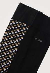 Pár čiernych ponožiek, jedna jednoliata s nápisom "BOSS" blízko manžety, druhá so vzorom hnedých a bielych geometrických tvarov na čiernom pozadí.
