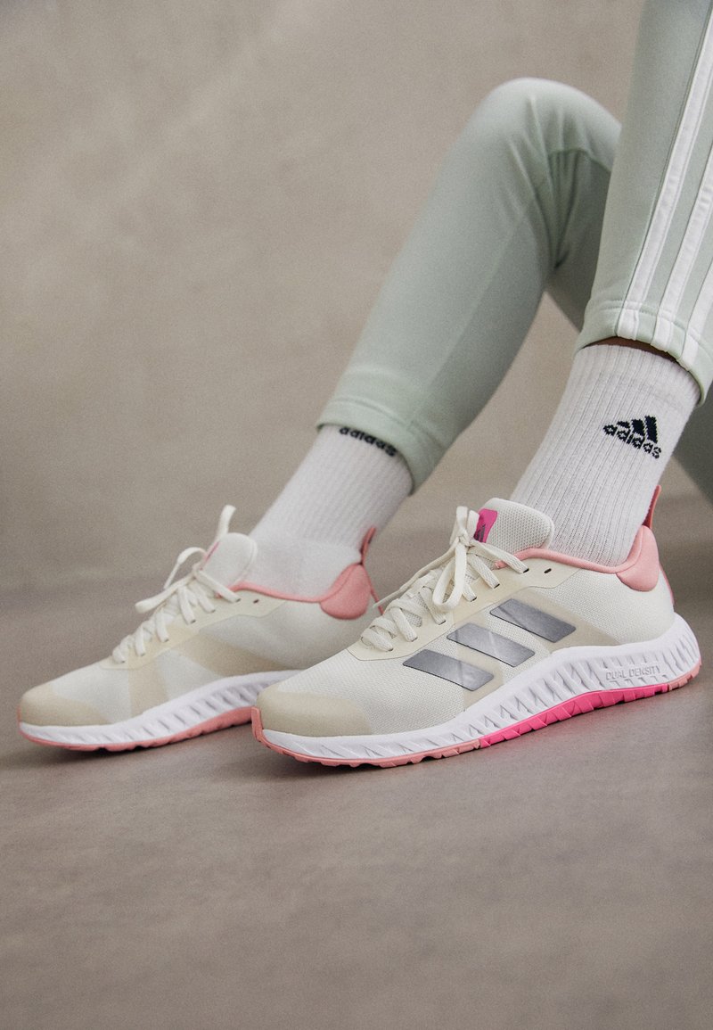 Zapatillas deportivas blancas con acentos en rosa claro y franjas plateadas en los lados. Parte superior de tejido texturizado con una suela de goma que presenta una suela rosa.