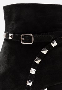 Stivaletto in suede nero con una fascia borchiata in metallo argento e un dettaglio di fibbia argentata alla caviglia. Texture liscia su tutta la superficie.