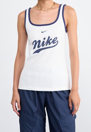 Kobieta w białym topie Nike z granatowym wykończeniem i granatowych spodniach bojówkach, pokazana od klatki piersiowej do ramion na jednolitym tle.