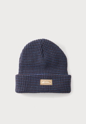 Bonnet tricoté bleu et marron avec revers plié et écusson rectangulaire beige avec logo à l'avant, sur fond clair uni.