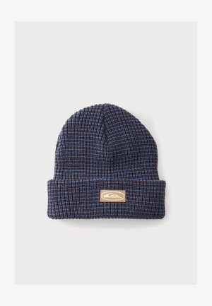 Bonnet tricoté bleu et marron avec revers plié et écusson rectangulaire beige avec logo à l'avant, sur fond clair uni.