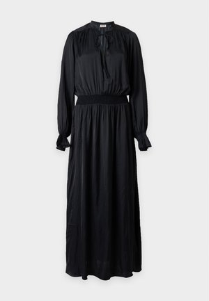 Femme aux cheveux bouclés portant une robe longue noire à manches longues avec une taille froncée et des talons noirs pointus, debout devant un fond uni.