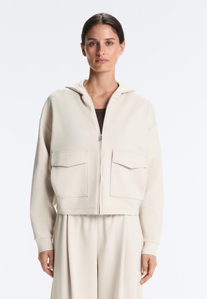 Vrouw draagt een lichtbeige zip-up hoodie met twee voorzakken met klep en bijpassende wijde broek, staand tegen een witte achtergrond.