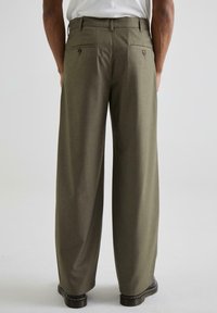 Pantaloni largi, de culoare verde măsliniu, dintr-un material texturat. Au două buzunare la spate cu nasturi și un croi drept, asortați cu pantofi negri.