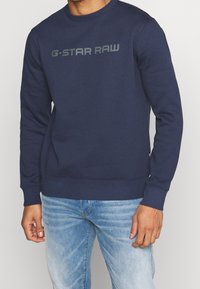 Man som bär en mörkblå G-Star Raw sweatshirt och ljusblå urtvättade jeans, stående mot en enfärgad bakgrund.