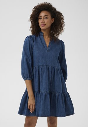 Kobieta z kręconymi włosami, ubrana w niebieską jeansową sukienkę warstwową z rękawami do łokcia i marszczonym kołnierzem, stojąca na jednolitym tle.