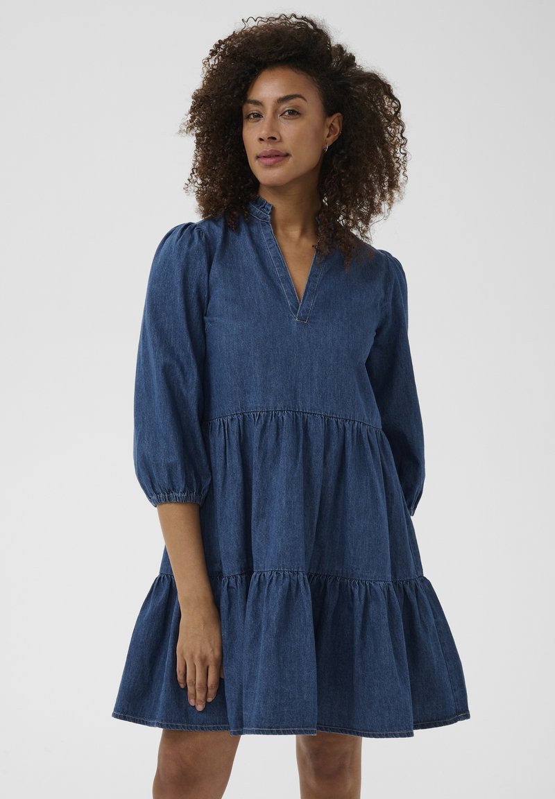 Femme aux cheveux bouclés portant une robe en jean bleue à volants avec des manches trois-quarts et un col à volants, debout devant un fond uni.