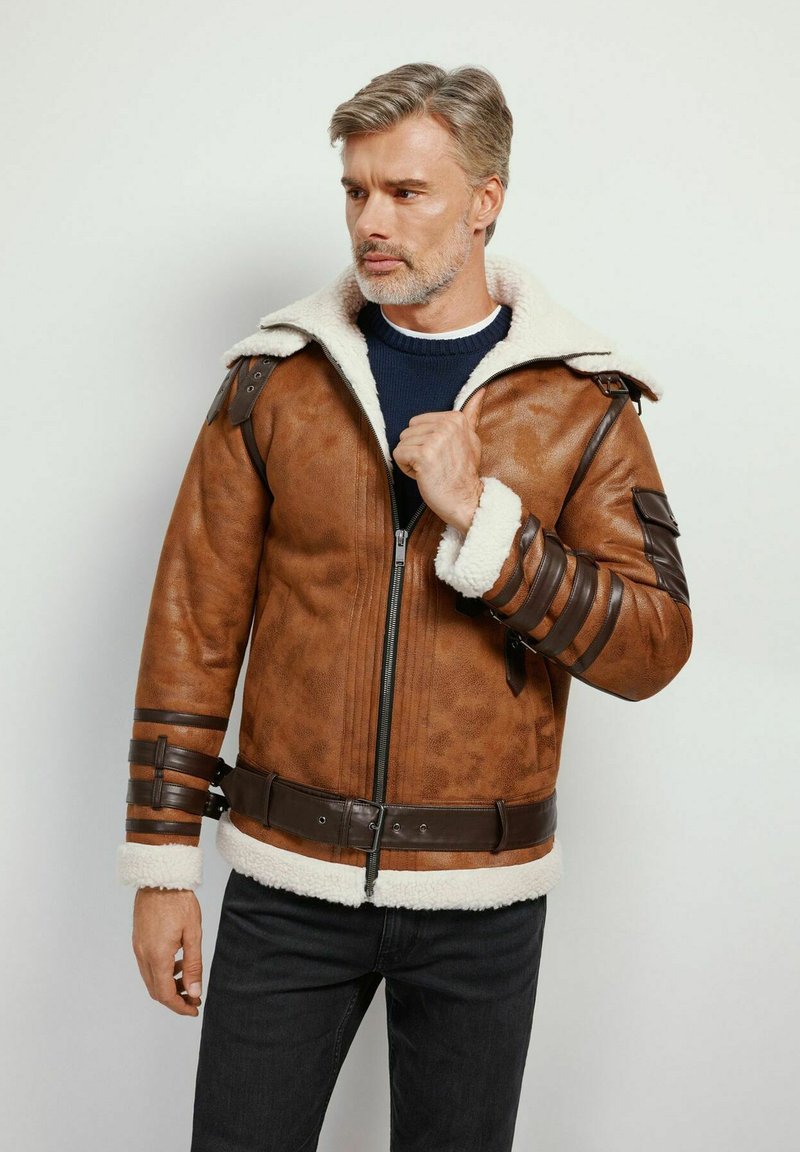 Giubbotto di pelle marrone con colletto in shearling, zip frontale e cinturini marroni. Caratterizzato da strisce di pelle orizzontali e una vita con cintura.