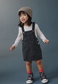 Robe salopette en denim noir avec boutons argentés, haut à manches longues blanc orné de pois rouges, chaussettes roses à volants, et baskets montantes noires à motifs.