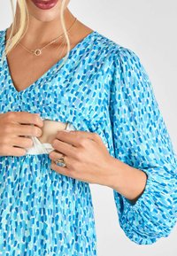 Blau gemustertes Kleid mit V-Ausschnitt aus leichtem Stoff. Lange Puffärmel und eine diskrete Öffnung für den Zugang. Goldener Ring an der Hand.