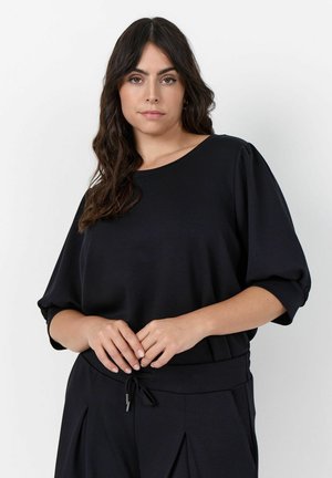 Frau mit langen, welligen dunklen Haaren, die ein lockeres schwarzes Oberteil und schwarze Hosen trägt, steht vor einem einfarbigen hellen Hintergrund.