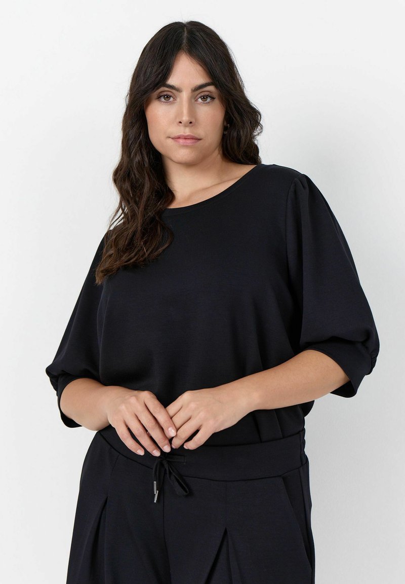 Femme aux longs cheveux ondulés et foncés, portant un haut noir ample et un pantalon noir, debout devant un fond clair uni.