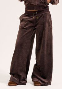 Wide-leg pants in dark brown, made of soft velvet. Features elastic waistband with drawstrings, large front pockets, and subtle logo detail.