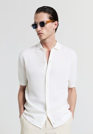 Chemise blanche texturée à manches courtes avec un devant à boutons et un col doux, accompagnée de lunettes de soleil à monture transparente avec des verres foncés et des accents marron.