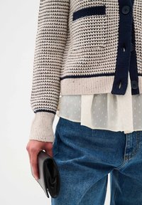 Knit cardigan in cream with navy stripes, featuring a pocket, paired with a sheer white blouse and blue denim jeans, holding a black wallet.