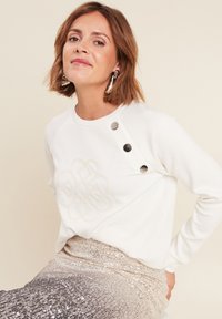 Weißer Pullover mit strukturiertem besticktem Design, ausgestattet mit silbernen Knöpfen an der Schulter. Kombiniert mit einem metallisch glitzernden Rock.