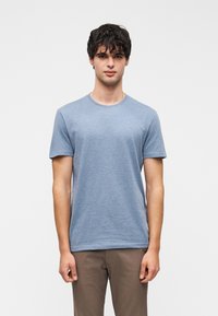 Νεαρός άνδρας με σκουρόχρωμα σγουρά μαλλιά, φορώντας ανοιχτό μπλε κοντομάνικη μπλούζα και καφέ παντελόνι, στέκεται μπροστά σε απλό λευκό φόντο.