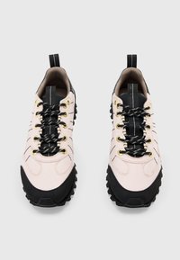Hellrosa Sportschuhe mit schwarzen Akzenten, strukturiertem Stoff, goldenen Ösen und dicken schwarzen Gummisohlen. Schwarze Schnürsenkel mit weißen Mustern.