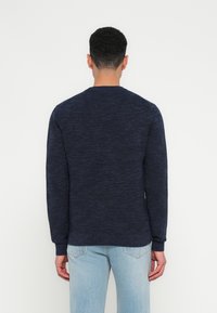 Man facing away wearing a dark blue knit sweater and light blue jeans against a plain white background.