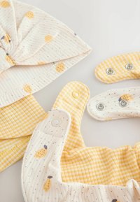 Gorro suave para bebé y babero de tela con estampados de cuadros amarillos y limones, con cierres de botones a presión sobre un fondo neutro.