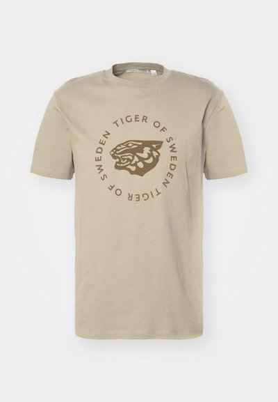 Bežs kokvilnas T-krekls ar apļveida grafiku, kurā attēlota tīģera galva un uzraksts "TIGER OF SWEDEN" brūnā krāsā. Īsās piedurknes un apaļš kakls.