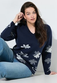 Granatowy sweter z dekoltem w serek z białymi kwiatowymi wzorami, charakteryzujący się luźnym krojem i miękką teksturą, zestawiony z jasnoniebieskimi dżinsami.