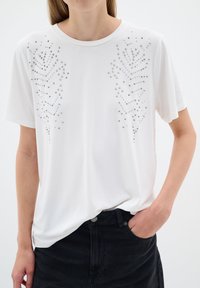Camiseta blanca de manga corta con adornos de tachuelas plateadas dispuestas en un patrón similar a plumas en el frente, con un corte relajado.