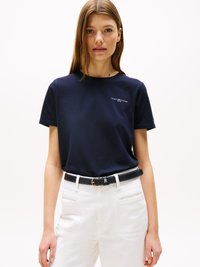 Žena ve tmavě modrém tričku Tommy Hilfiger zastrčeném do bílých kalhot s vysokým pasem a černým opaskem, stojící na jednobarevném pozadí.