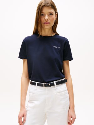 Woman wearing a navy blue Tommy Hilfiger t-shirt tucked into high-waisted white pants with a black belt, standing against a plain background.
