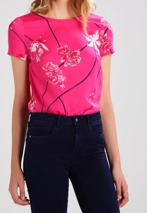 Vrouw draagt een felroze blouse met korte mouwen en bloemenpatroon, ingestopt in donkerblauwe spijkerbroek.