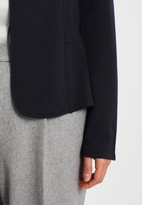 Laivastonsininen neulottu bleiseri, jossa on sileä pinta ja jäsennelty malli, yhdistettynä vaaleanharmaisiin villahousuihin. Ominaisuuksiin kuuluu pyöristetty helma ja pitkät hihat.