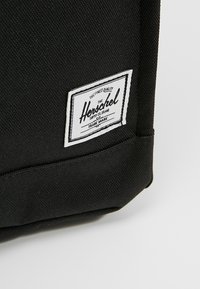Gros plan du coin d’un sac en tissu noir avec une étiquette blanche rectangulaire de la marque Herschel cousue dessus.
