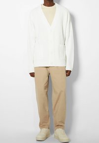 Vit ribbad cardigan med V-hals och två framfickor, bärs över en beige topp och matchas med beiga byxor och ljusa sneakers.