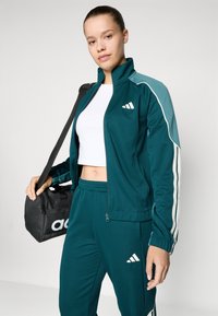 Tamsiai mėlynas treningas su baltais dryžiais, su užtrauktuku užsegama striukė ir kelnes su elastingu liemeniu. Juodas sportinis krepšys su baltais logotipo akcentais.