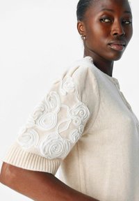 Kvinna som bär en beige stickad kortärmad tröja med intrikata vita blommönster broderade på ärmen, blickande åt sidan.