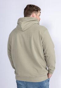 Felpa color verde oliva chiaro con una texture morbida, dotata di tasca a marsupio frontale, polsini a coste e cappuccio con coulisse, indossata con jeans blu.